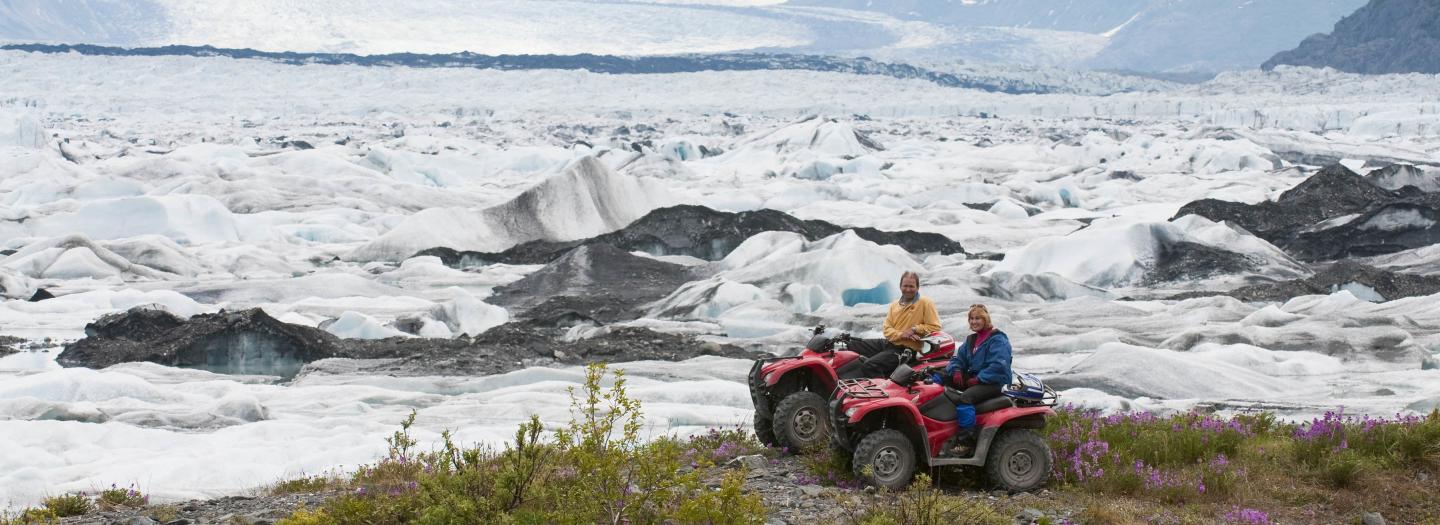 Two people on red ATVs explore a vast, icy glacier landscape with snow-capped mountains in the distance in Mat-Su, Alaska