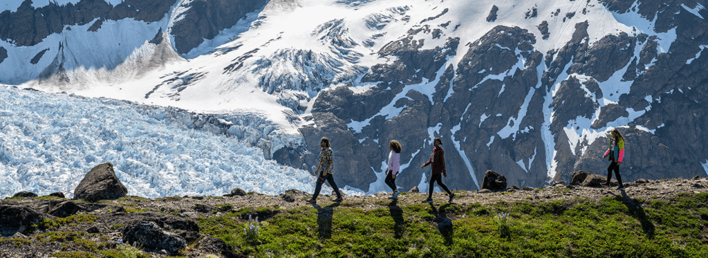 Four hikers traverse a grassy ridge with a breathtaking view of a snow-capped mountain and glacier in the distance in Mat-Su, Alaska
