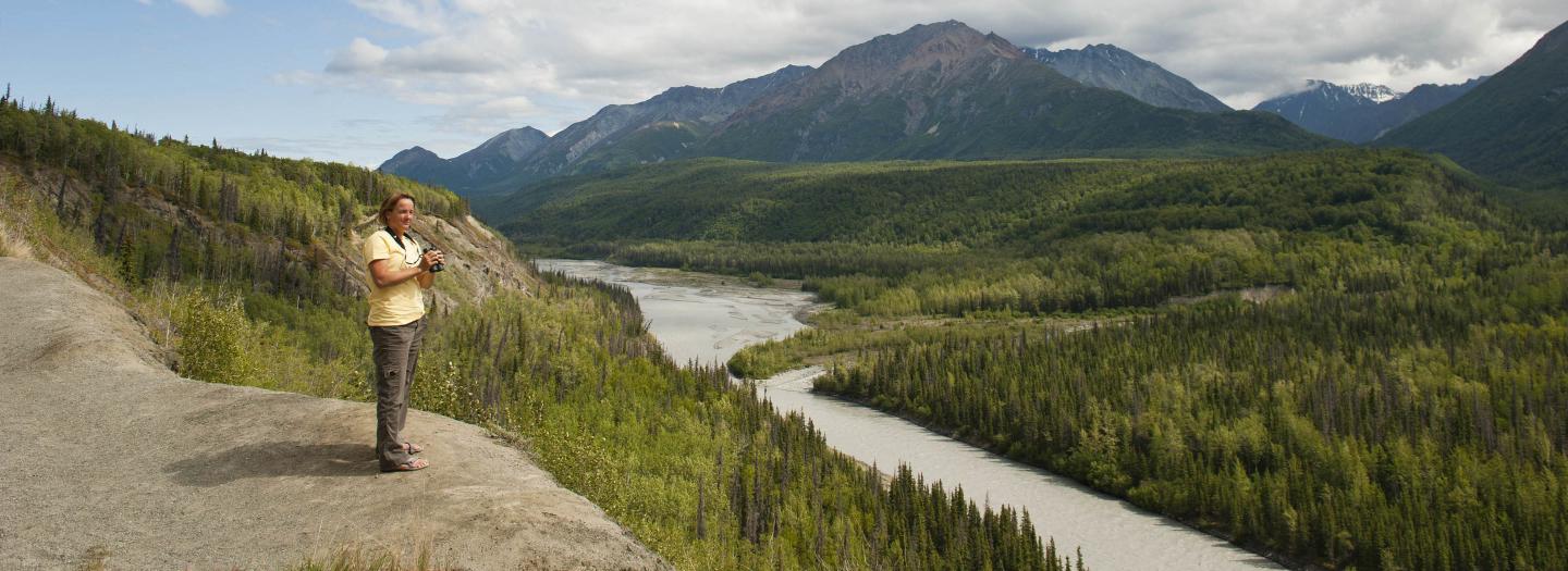 A person stands on a rocky outcrop overlooking a vast, winding river surrounded by lush green forests and towering mountains under a partly cloudy sky in Mat-Su, Alaska