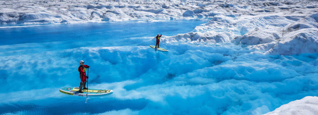 Three paddleboarders navigate through vibrant blue glacial waters surrounded by towering ice formations in Mat-Su, Alaska