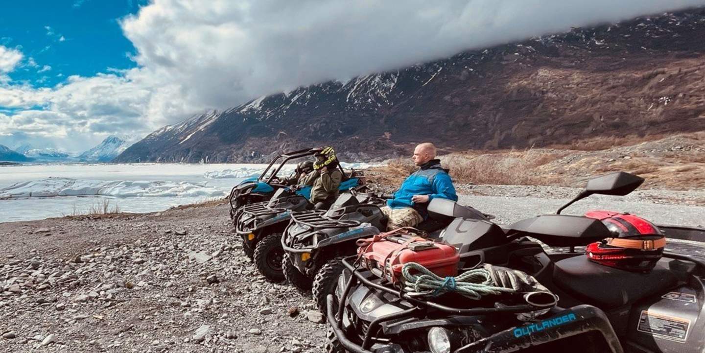 A group of ATVs parked on a rocky shore near a frozen lake, with snow-capped mountains in the background in Mat-Su, Alaska