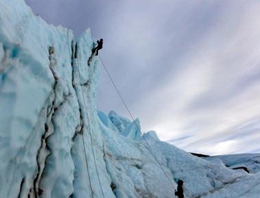 Two climbers ascend a towering ice cliff, secured by ropes, under a cloudy sky in Mat-Su, Alaska
