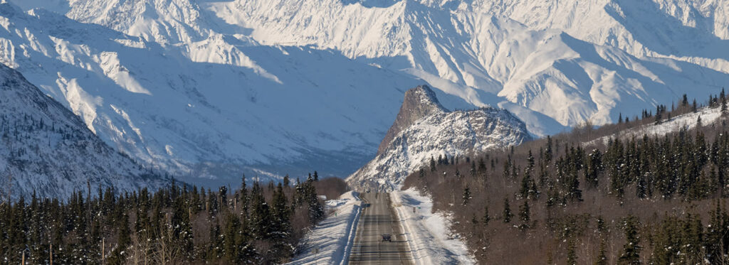 A winding road cuts through a snowy mountain landscape, flanked by towering peaks and evergreen forests in Mat-Su, Alaska