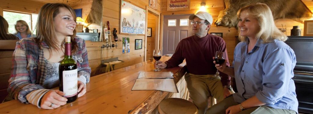 Three people enjoying wine at a rustic wooden bar in a cozy cabin setting in Mat-Su, Alaska