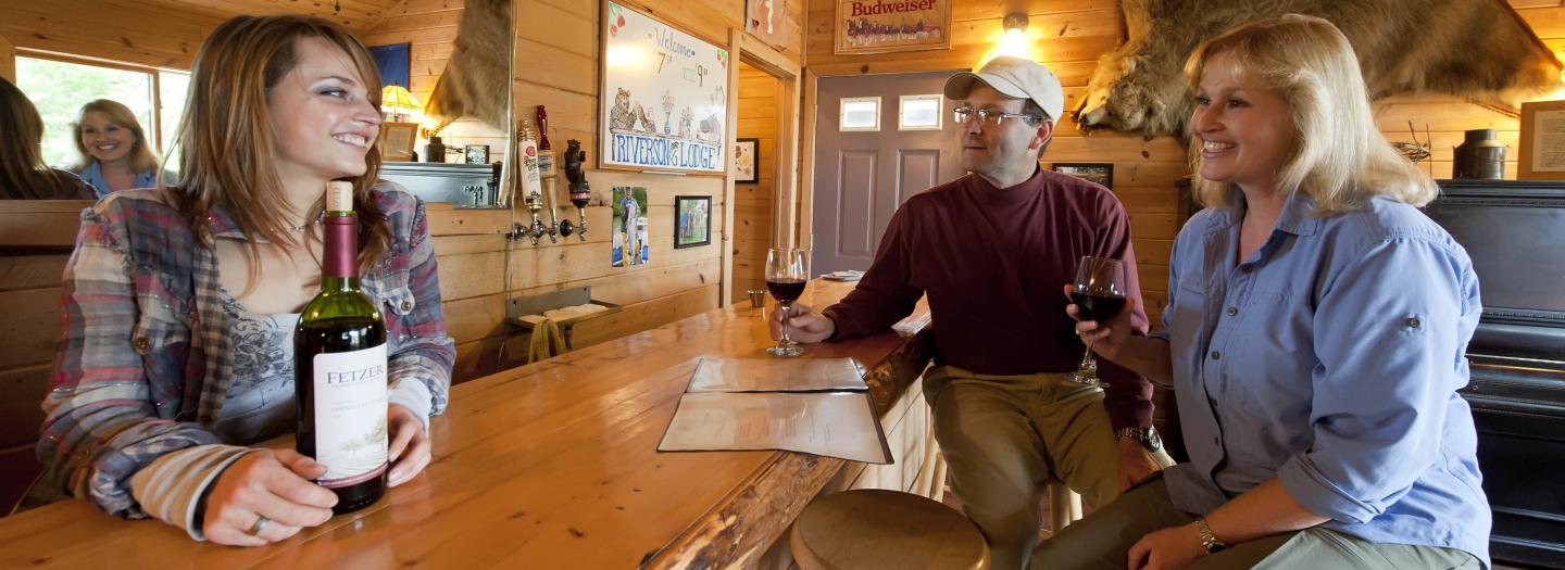Three people enjoying wine at a rustic wooden bar in a cozy cabin setting in Mat-Su, Alaska