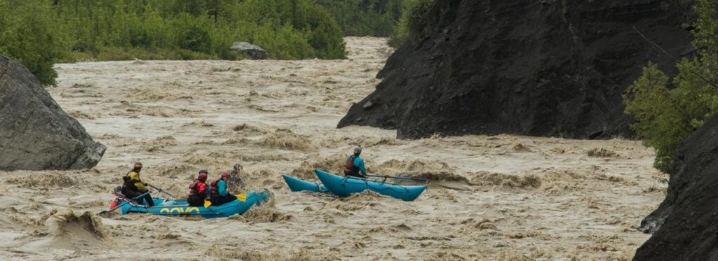 Four kayakers navigate through rough, muddy waters in a narrow canyon, surrounded by steep rock walls and lush greenery in Mat-Su, Alaska