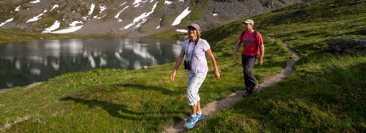 Two hikers walk along a mountain trail near a serene alpine lake surrounded by snow-capped peaks in Mat-Su, Alaska
