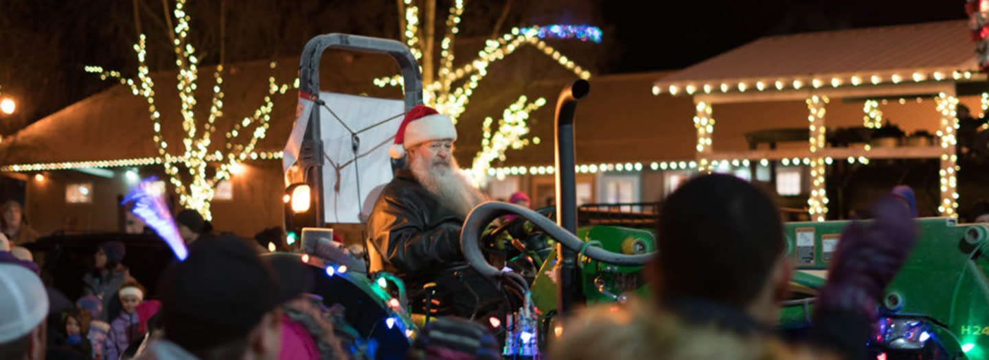 A festive parade scene at night featuring Santa Claus on a tractor, surrounded by cheering crowds and illuminated by bright Christmas lights in Mat-Su, Alaska