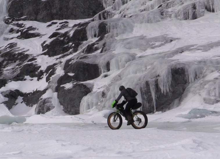 A cyclist rides a fat-tire bike across a snowy landscape with icy cliffs in the background in Mat-Su, Alaska
