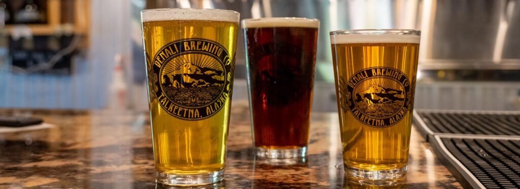 Three glasses of beer with different colors and head sizes sit on a bar counter in Mat-Su, Alaska