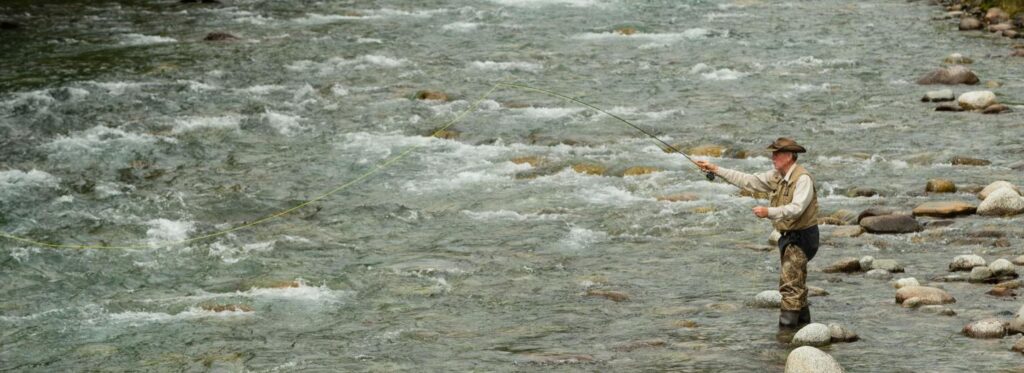 A person wearing waders stands in a fast-flowing river, casting a fishing line into the water in Mat-Su, Alaska