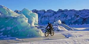 A cyclist rides across a frozen landscape with towering blue ice formations in the background in Mat-Su, Alaska