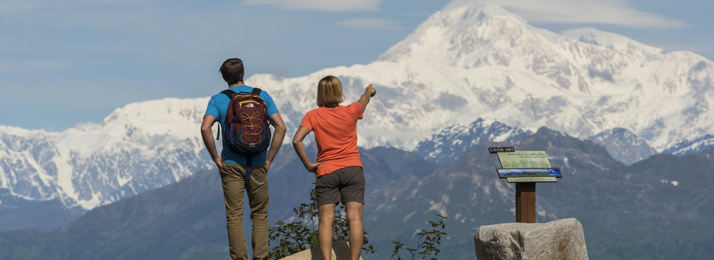 A couple stands on a rocky outcrop, admiring a majestic snow-capped mountain range under a clear blue sky in Mat-Su, Alaska
