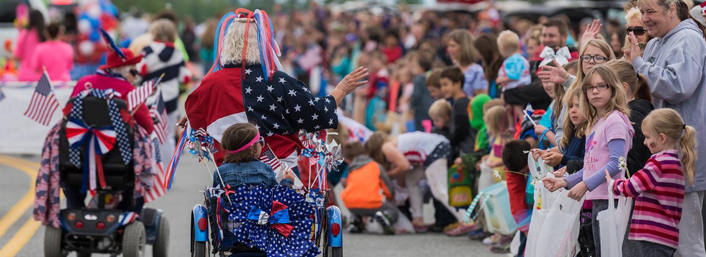 A vibrant Fourth of July parade featuring participants in wheelchairs adorned with patriotic decorations, waving to a cheering crowd in Mat-Su, Alaska