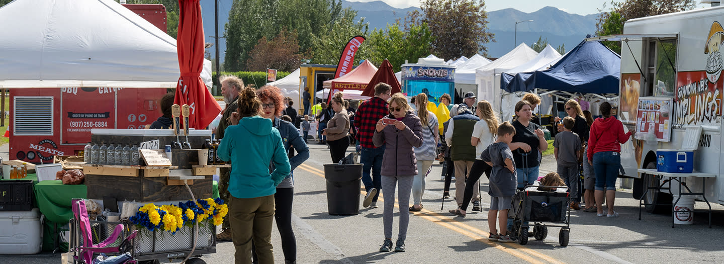 A bustling outdoor market with colorful tents, food trucks, and a lively crowd of shoppers and vendors in Mat-Su, Alaska