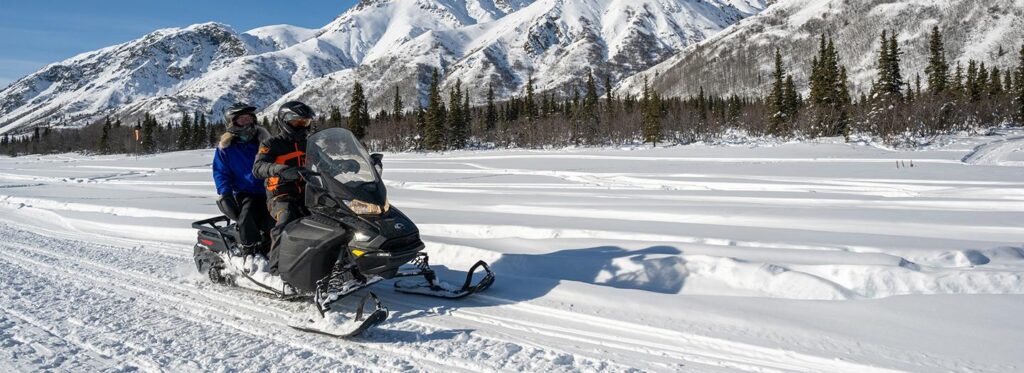 Two people riding a snowmobile across a snowy landscape with mountains in the background in Mat-Su, Alaska