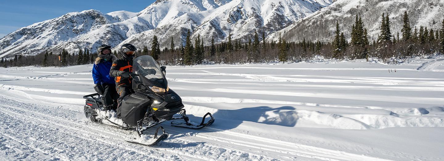 Two people riding a snowmobile across a snowy landscape with mountains in the background in Mat-Su, Alaska