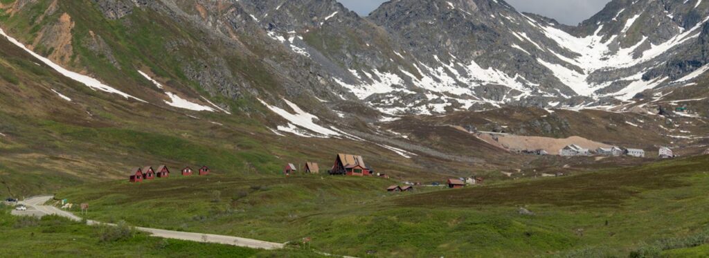 A panoramic view of a lush green valley nestled between towering snow-capped mountains with scattered wooden cabins in Mat-Su, Alaska