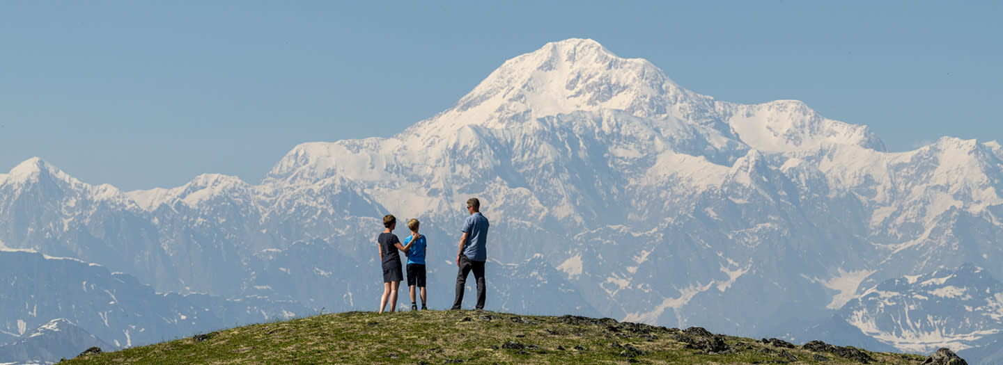 A family of four stands on a grassy hill, gazing at the majestic snow-capped mountains in the distance under a clear blue sky in Mat-Su, Alaska