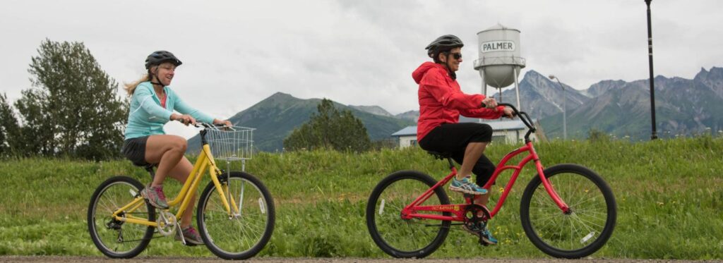 Two cyclists, one in a blue shirt and the other in a red jacket, ride their yellow and red bicycles on a grassy path with mountains in the background in Mat-Su, Alaska
