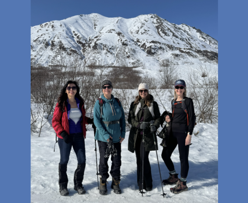Four women dressed in winter gear stand together on a snowy landscape, with a majestic snow-capped mountain in the background in Mat-Su, Alaska