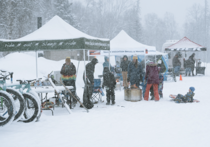 A snowy outdoor event with people gathered around tents, enjoying winter activities and warm drinks in Mat-Su, Alaska