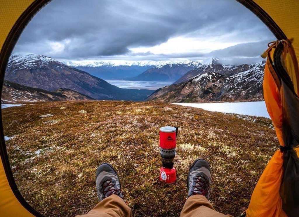 A hiker's perspective from inside a yellow tent, looking out at a breathtaking mountain landscape with snow-capped peaks, a serene lake, and a red camping stove in the foreground in Mat-Su, Alaska