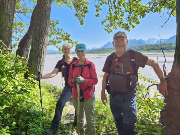 Three hikers pose for a photo in a lush forest near a river, with mountains in the distance in Mat-Su, Alaska