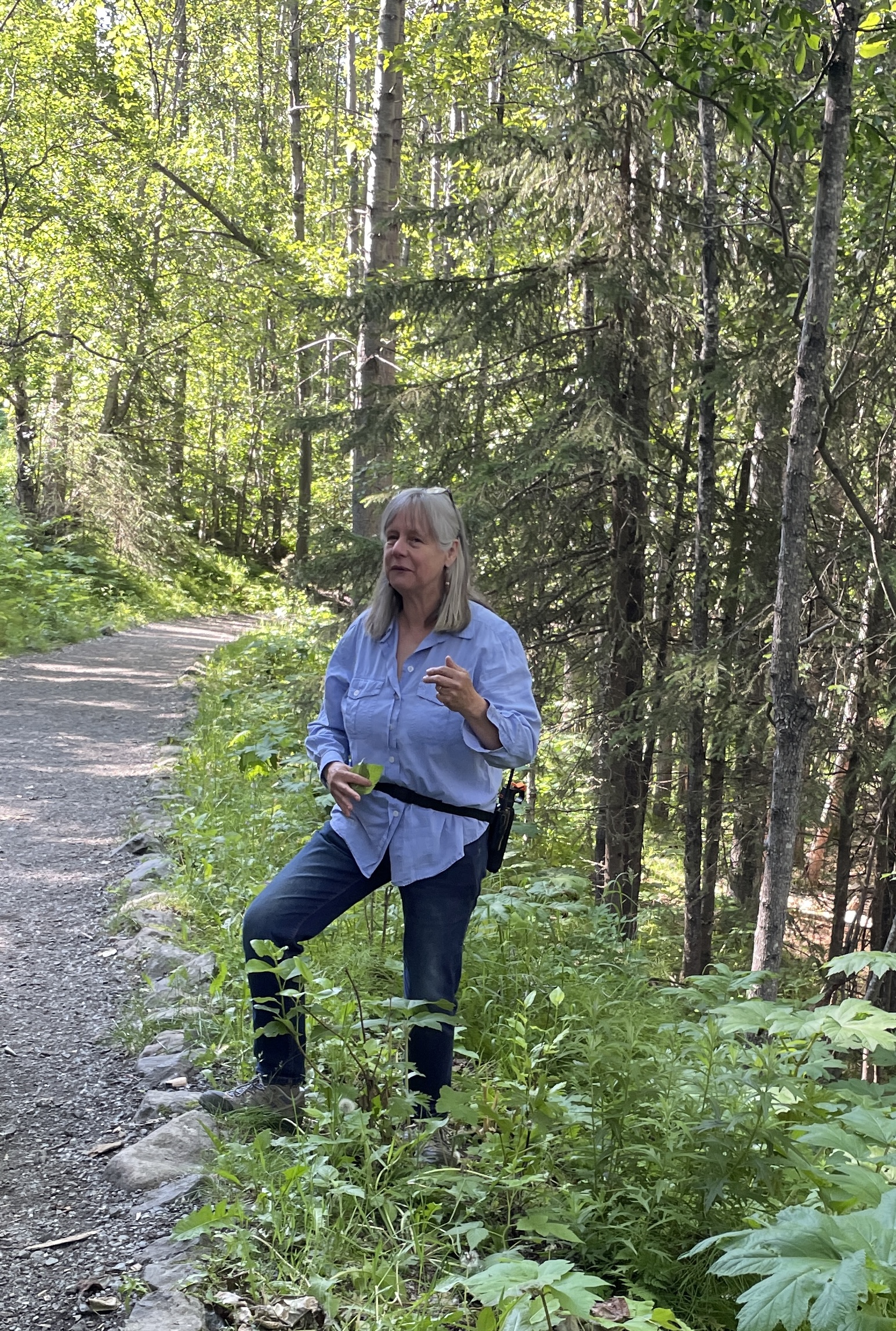 A woman in a blue shirt stands on a dirt path in a lush forest, surrounded by tall trees and green undergrowth in Mat-Su, Alaska