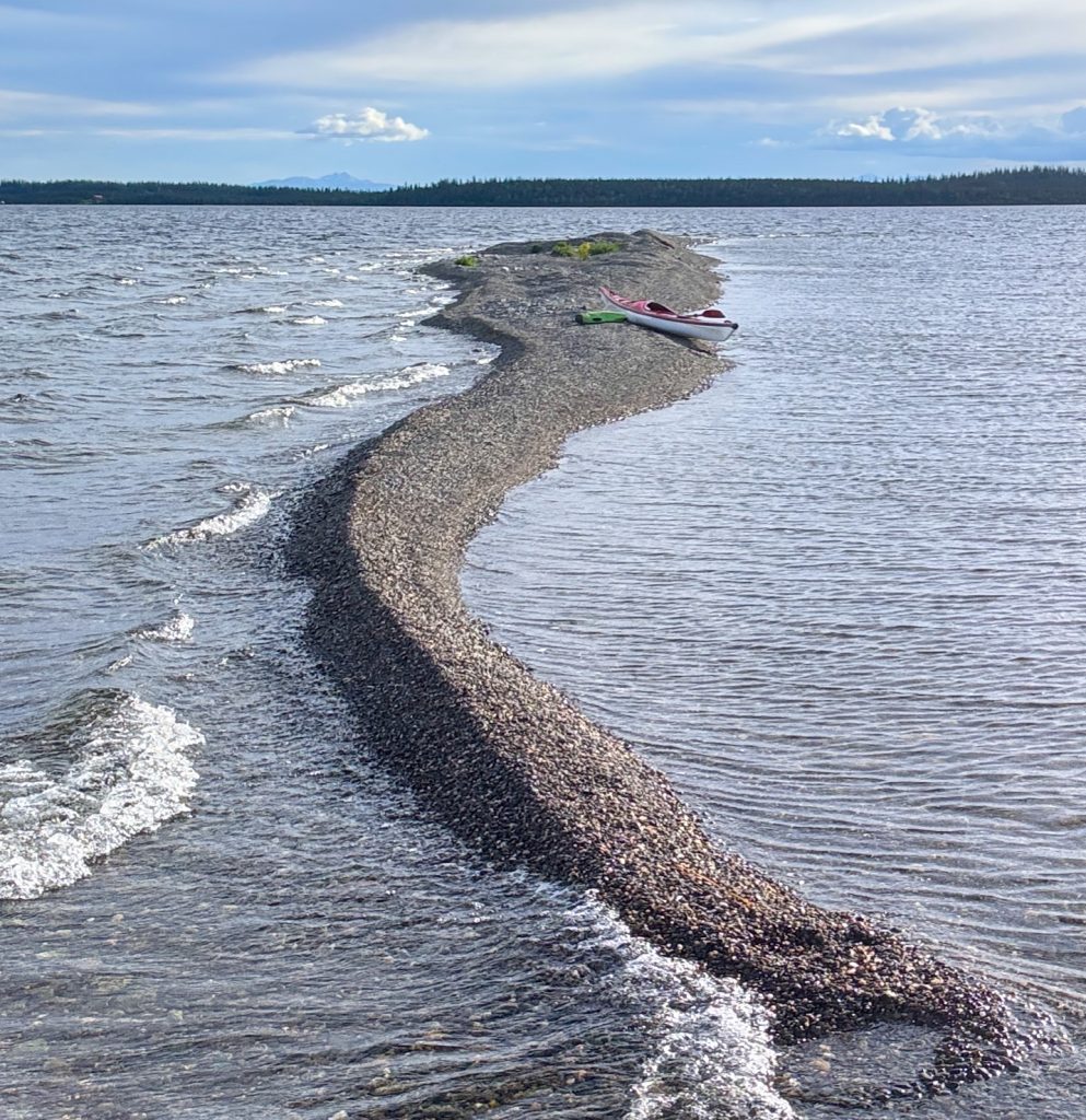 A winding pebble beach leads to a small island with a kayak on a calm lake in Mat-Su, Alaska