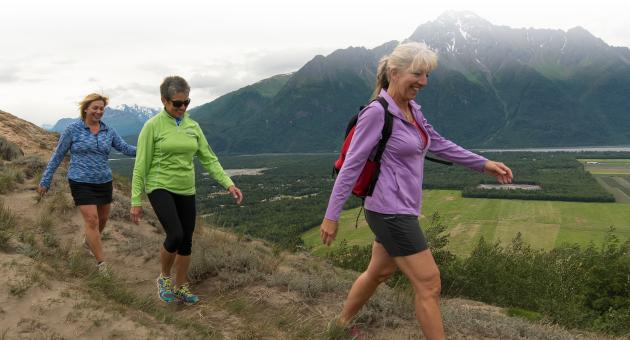 Three women hike along a mountain trail with a scenic view of valleys and distant mountains in Mat-Su, Alaska