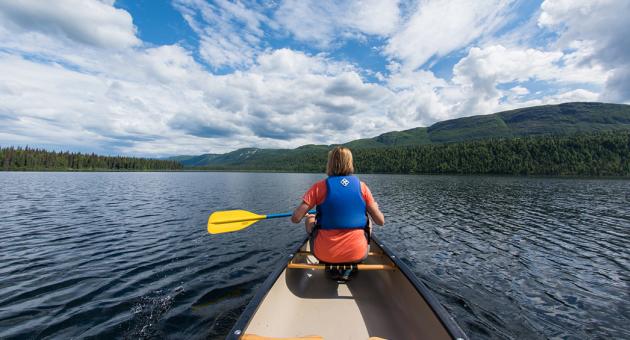 A person in a red shirt paddles a canoe on a serene lake, surrounded by lush green hills and a partly cloudy sky in Mat-Su, Alaska