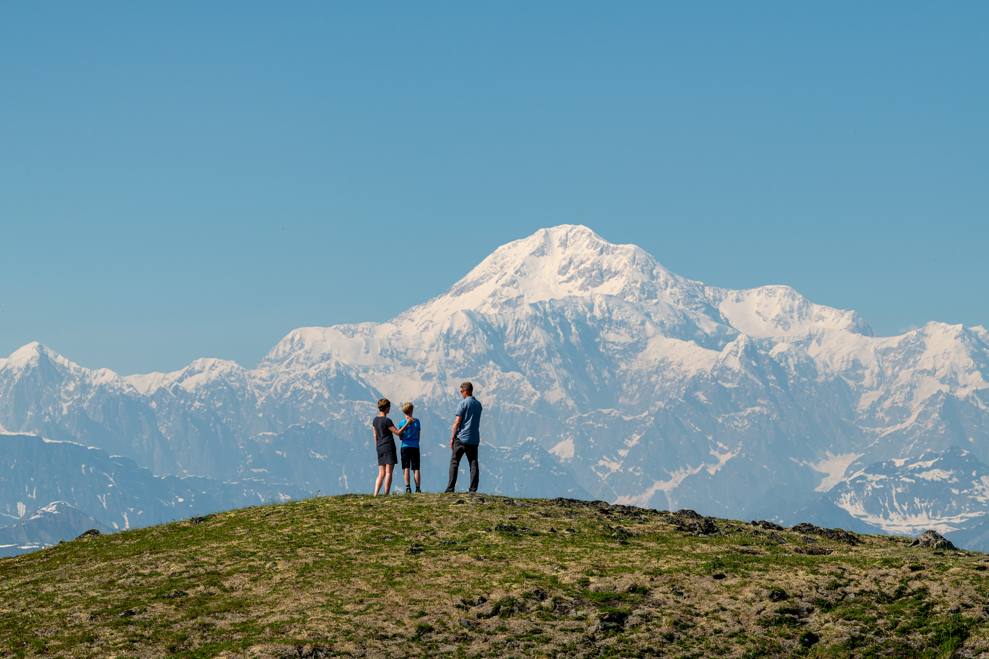Three people stand on a grassy hill, gazing at a majestic snow-capped mountain under a clear blue sky in Mat-Su, Alaska