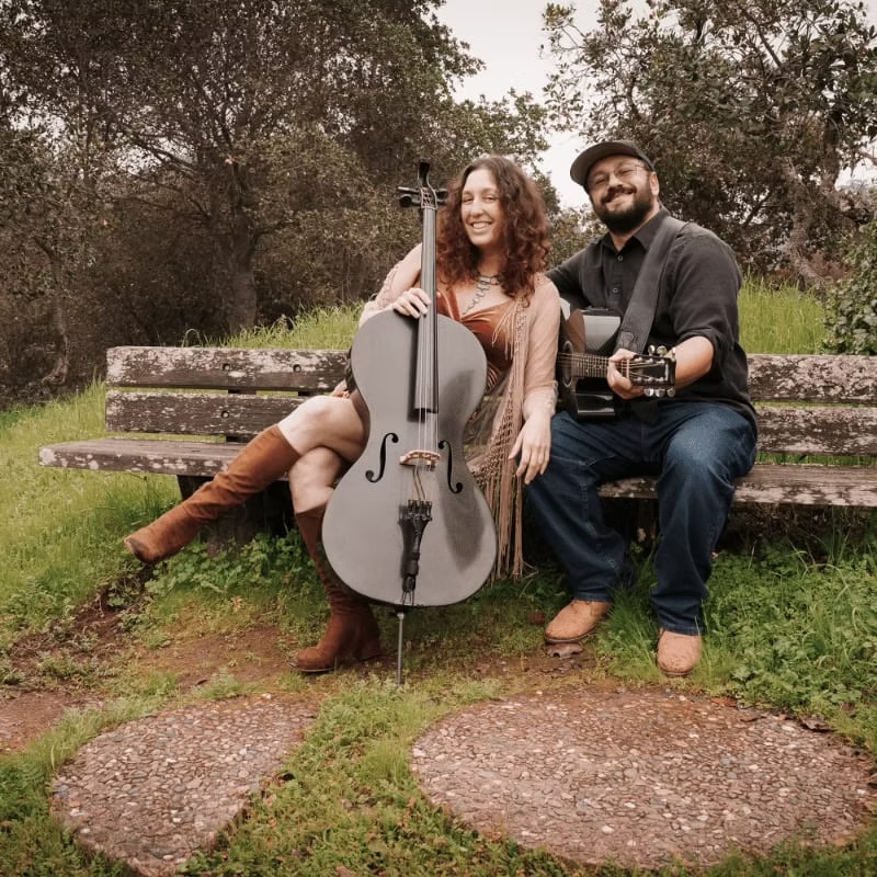 A couple sits on a bench with a cello and guitar, surrounded by nature in Mat-Su, Alaska