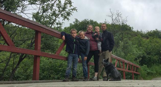 A family of five, including a baby, stands on a wooden bridge with a red railing, accompanied by a black and white dog in Mat-Su, Alaska