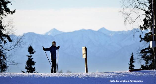 A skier stands atop a snowy hill, holding ski poles, with a signpost nearby and majestic mountains in the distance in Mat-Su, Alaska
