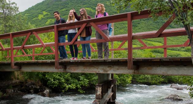 Four friends enjoy a scenic view from a wooden bridge over a rushing river, surrounded by lush greenery in Mat-Su, Alaska