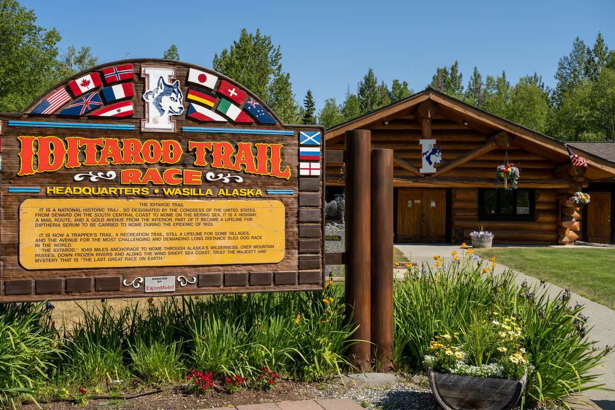 A rustic wooden sign for the Iditarod Trail Race with colorful flags and a husky logo stands in front of a log cabin headquarters surrounded by lush greenery and flowers in Mat-Su, Alaska