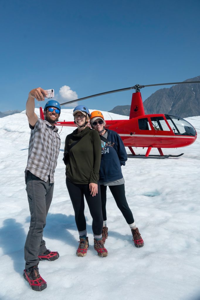 Three adventurers in colorful helmets and winter gear take a selfie with a red helicopter on a snowy glacier under a clear blue sky in Mat-Su, Alaska