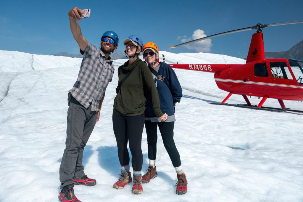 Three adventurers in winter gear take a selfie with a red helicopter on a snowy landscape in Mat-Su, Alaska