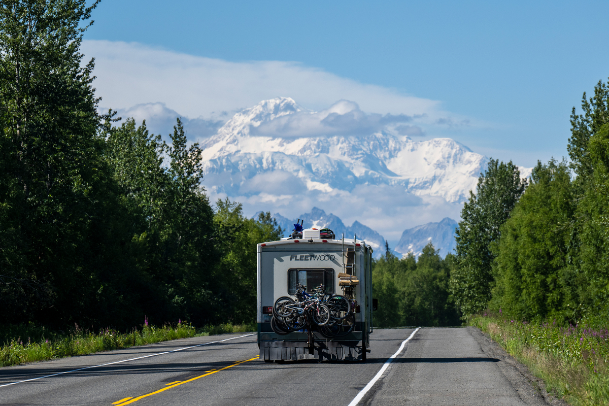 A recreational vehicle with bicycles attached travels down a scenic highway flanked by lush green trees, with majestic snow-capped mountains in the distance under a clear blue sky in Mat-Su, Alaska