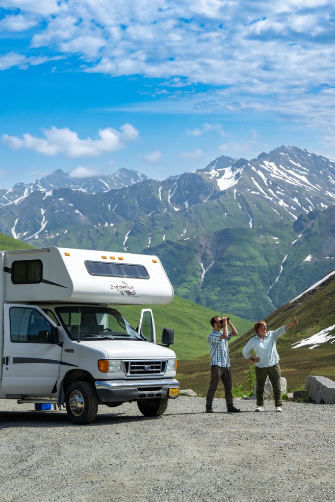 A couple stands near a white camper van, gazing at the majestic snow-capped mountains in the distance under a clear blue sky in Mat-Su, Alaska