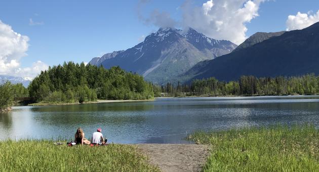 A serene lakeside scene with two people enjoying a picnic on the grassy shore, framed by majestic snow-capped mountains and lush green forests in Mat-Su, Alaska