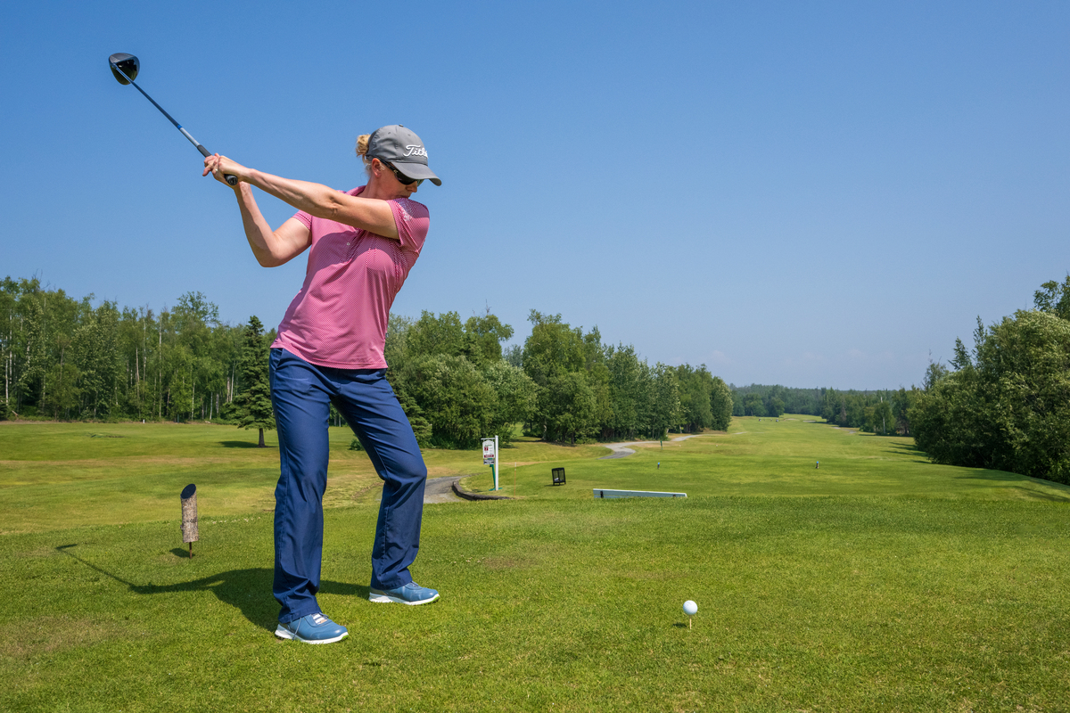 A golfer in a pink shirt and blue pants is swinging a club on a sunny day at a golf course in Mat-Su, Alaska