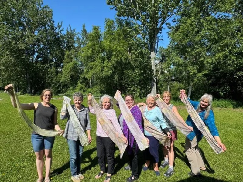 A group of seven women joyfully pose outdoors, each holding a long, colorful scarf that they wave in the air in Mat-Su, Alaska
