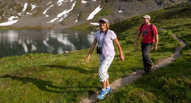 A couple enjoys a hike along a mountain lake trail, surrounded by snow-capped peaks and lush greenery in Mat-Su, Alaska