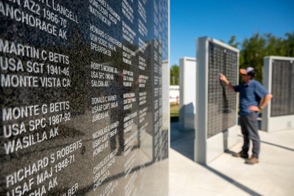 A man stands before a wall of engraved names, likely a memorial, under a clear blue sky in Mat-Su, Alaska