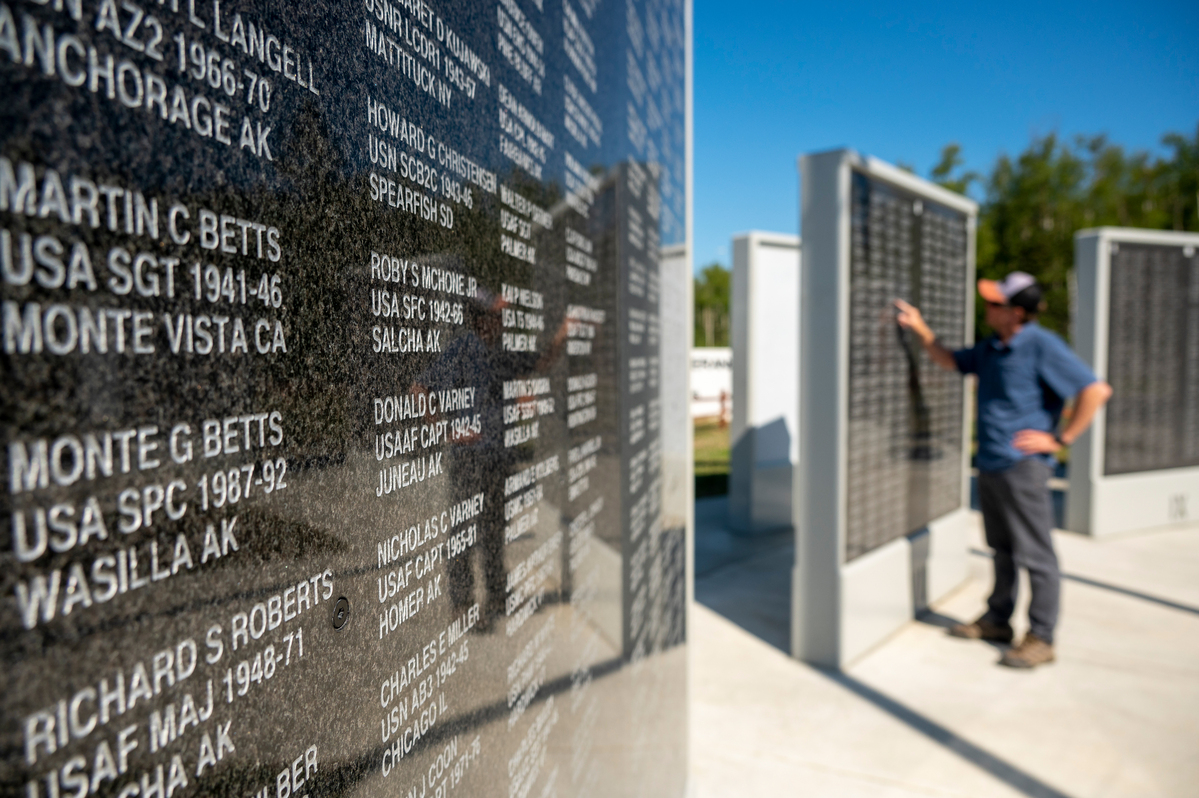 A man stands before a wall of engraved names, likely a memorial, under a clear blue sky in Mat-Su, Alaska