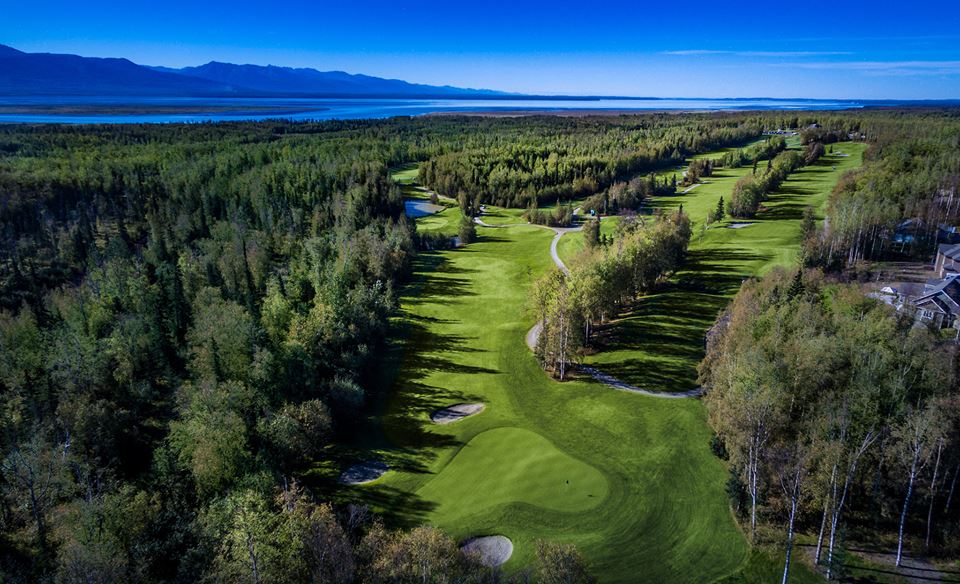 An aerial view of a lush green golf course winding through a dense forest with a distant mountain range and clear blue sky in Mat-Su, Alaska