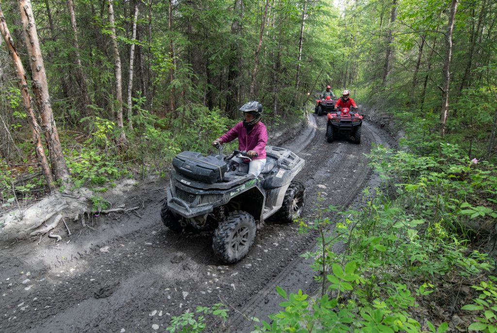 Four ATV riders navigate a muddy trail through a lush forest, kicking up dirt and enjoying the outdoor adventure in Mat-Su, Alaska